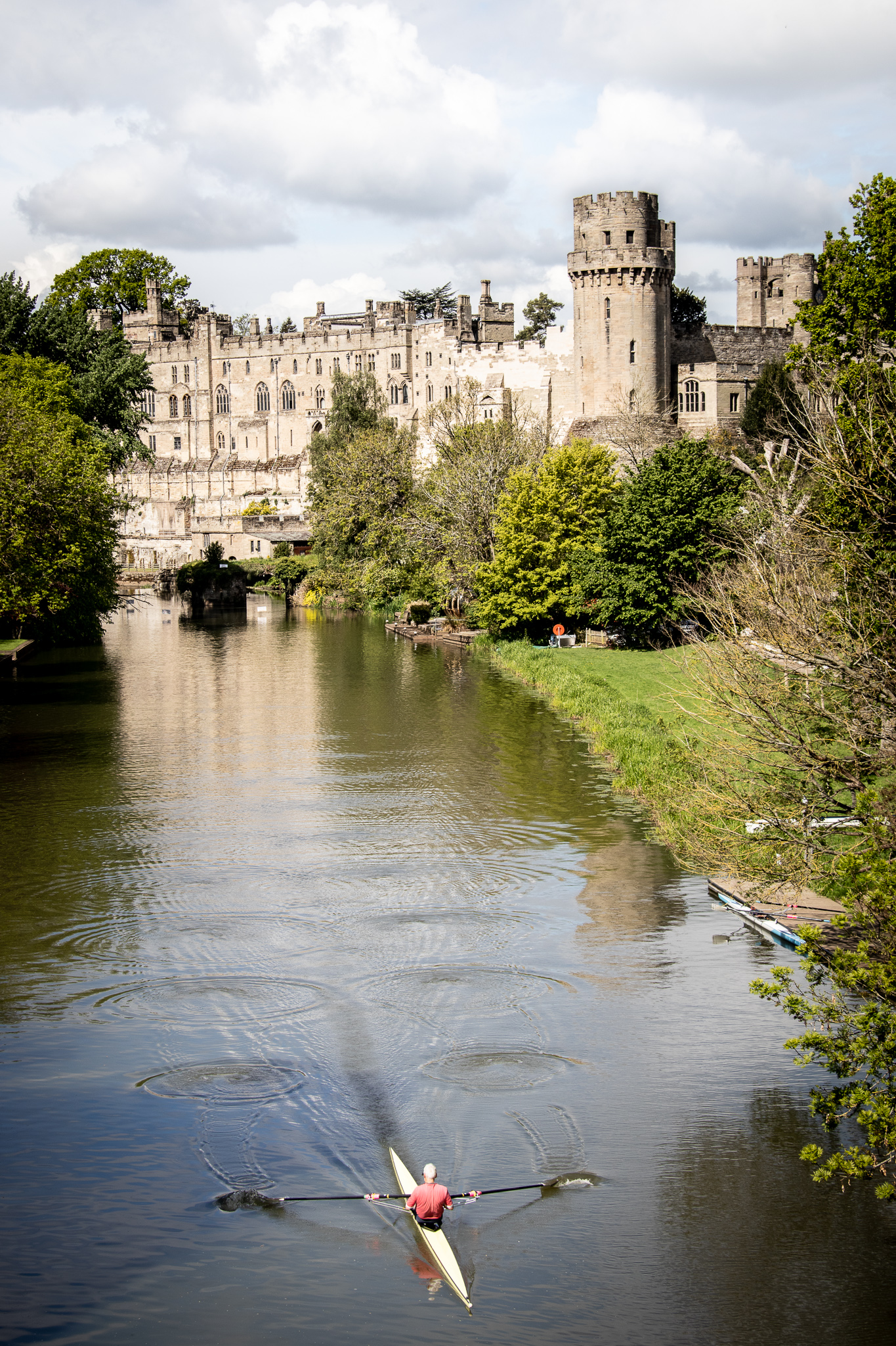 A view of Warwick Castle from Castle Bridge on the River Avon. A man is rowing down the River Avon and ripples show his path down the river. 