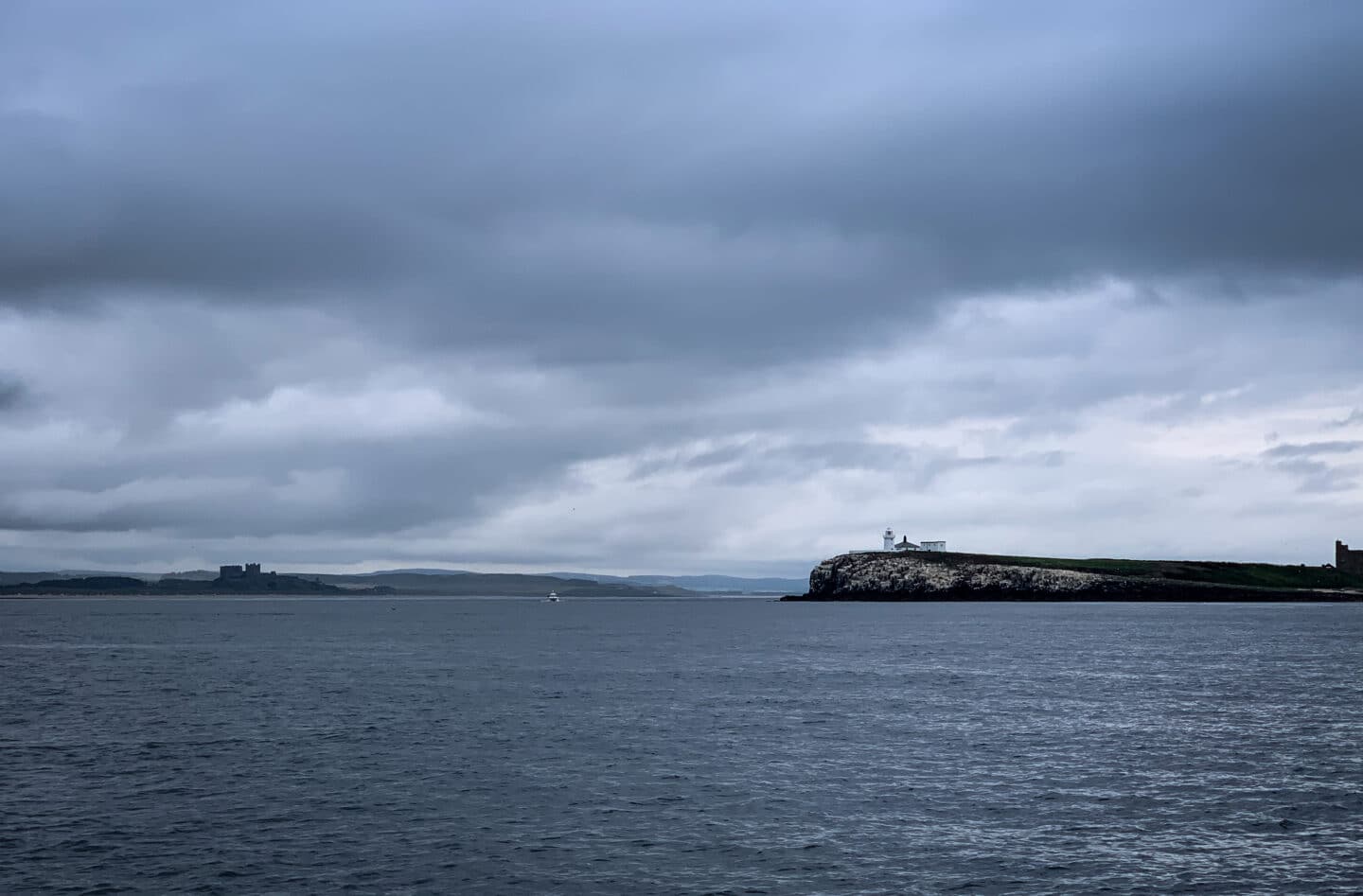 A view from our Farne Island tour boat - on the horizon you can see the Inner Farne and Its lighthouse on the right, and Bamburgh Castle in the distance. The skies are moody and grey. 