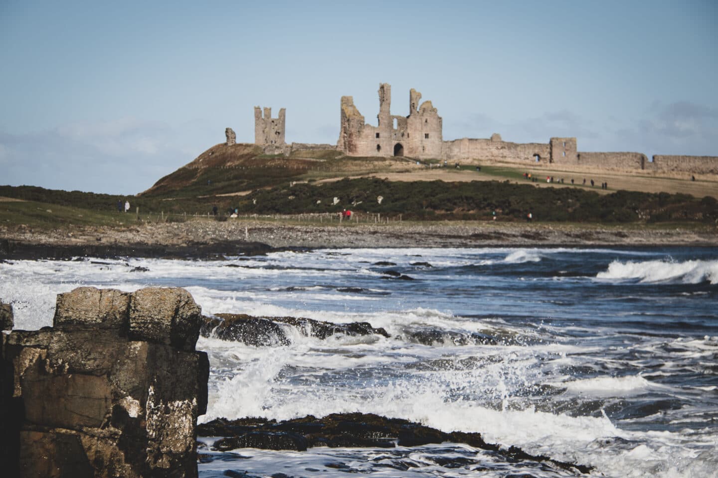 A view of Dunstanburgh Castle over the North Sea Coast. Waves are crashing into the rocks in the foreground. 